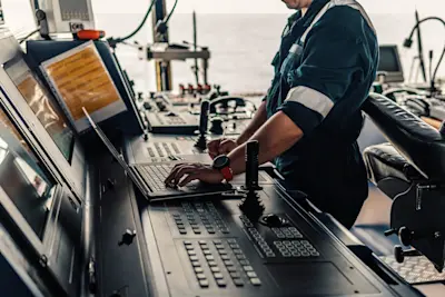 Photo of a person in a boiler suit on the bridge of a ship, standing up and typing on a laptop with one hand.